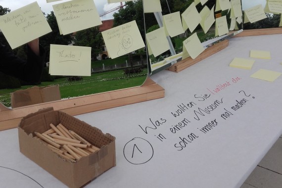 Ideen-Tisch mit Frage auf Papiertischdecke &quot;Was wolltest du schon immer mal im Museum machen?&quot;, danebem Box mit Bleistiften, Aufsteller mit PostIds