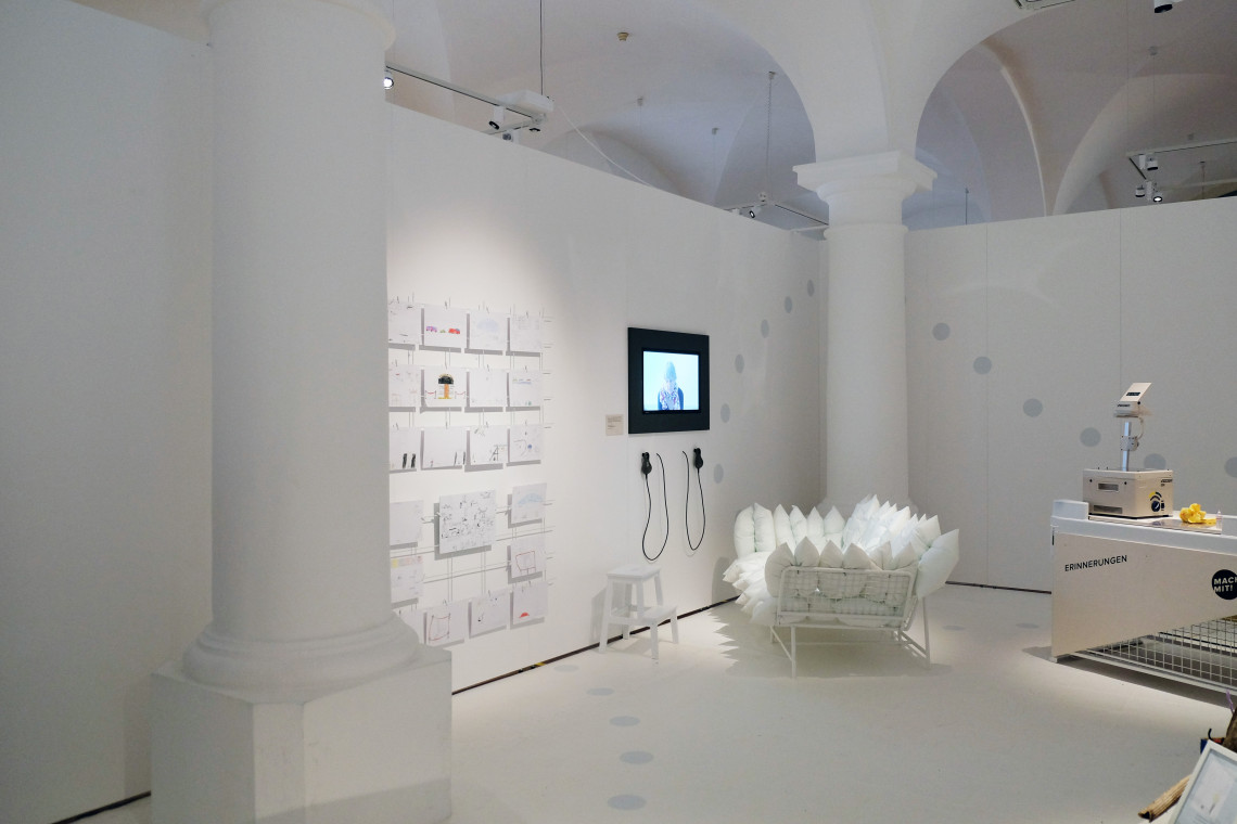 Ganz in Weiß gestalteter Ausstellungsraum mit Zeichnungen an der Wand, Viedeoscree, Sofa übersäht mit weißen Kissen
