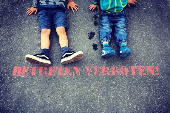Kinder sitzend auf Asphalt. Vor Füßen steht auf dem Asphalt in roter Schrift: Betreten verboten!