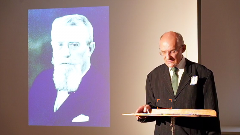 Dr. Gerhard Hölzle am Rednerpult, im Hintergrund ein Foto von Bernhard Stirnweiß an die Wand projeziert