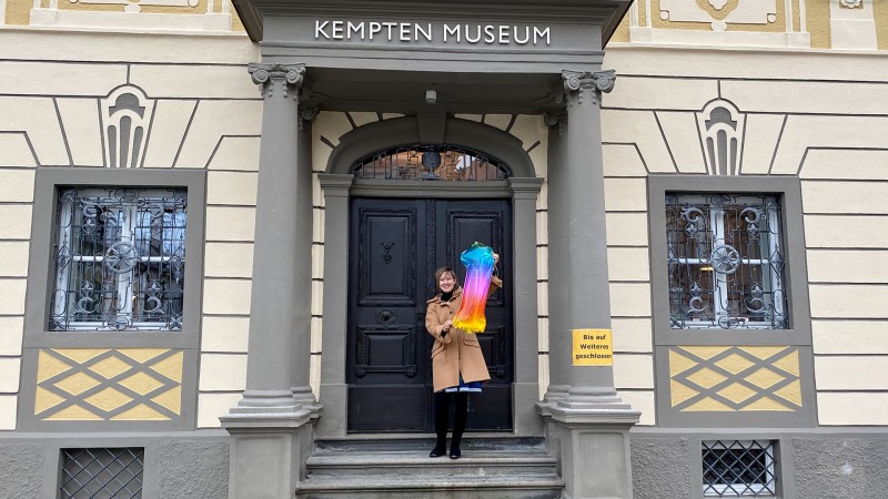 Museumsleiterin Christine Müller Horn hält großen Folienballon (Ziffer 1) in Regenbogenfarben vor Eingangstür Kempten-Museum
