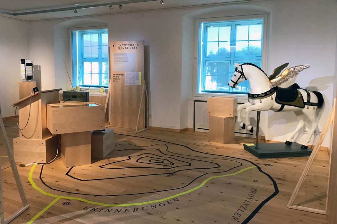 Ausstellungsraum mit Ausstellungsmöbeln in hellem Holz, Linien auf Boden, Hörstation, lebensgroßer Pegasus-Figur