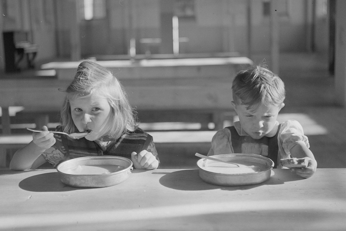 Schwarz-Weiß-Bild: ein Mädchen und ein Junge (ca. sieben und fünf Jahre alt) löffeln Suppe.