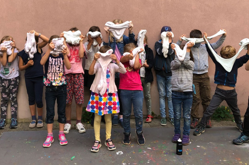 Kinder vor Mauer, die selbst hergestelltem Leucht-Schleim vor ihre Gesichter halten