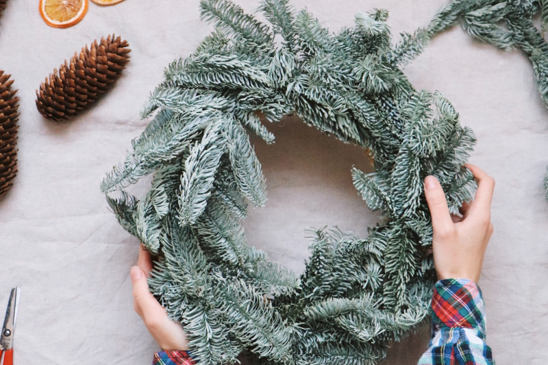 Hände greifen Adventskranz, daneben Tannenzapfen, Schere, Orangenschalen